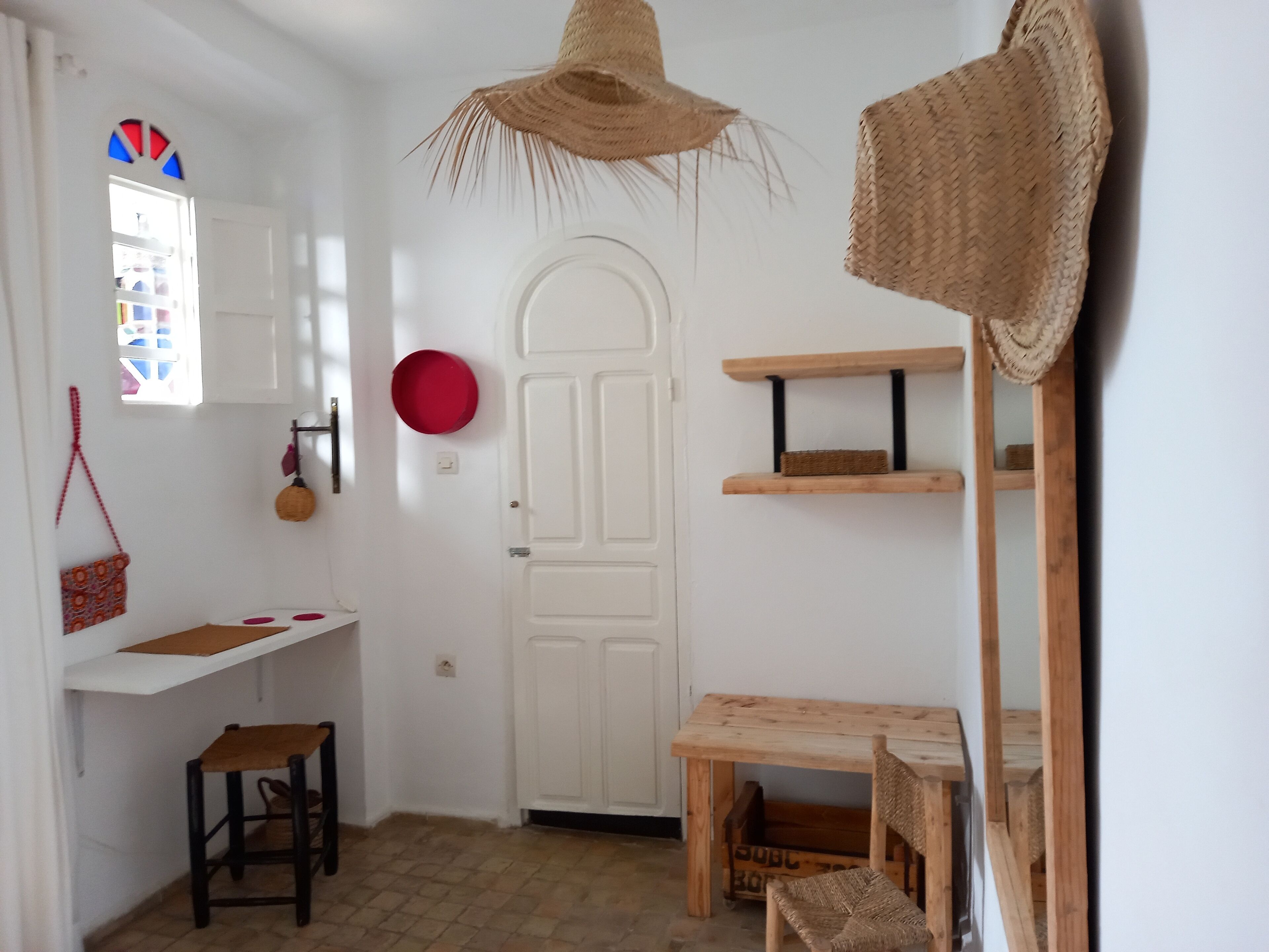A room with a white door, a small wooden table, a chair, a hanging basket, and a red bowl on the wall.