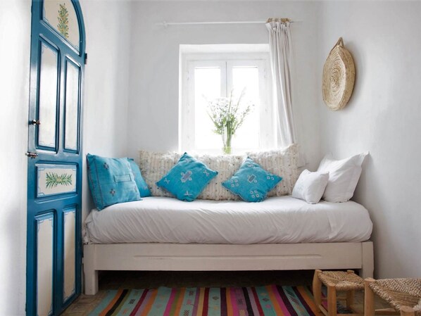 A bedroom with a white bed, blue pillows, a window with white curtains, and a woven wall hanging. - Hotel Emeraude Essaouira (Essaouira)
