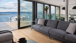 Un salon moderne avec un canapé gris, une petite table et des chaises. Une grande porte-fenêtre coulissante donne accès à un balcon d’où l’on peut admirer la mer et les montagnes.