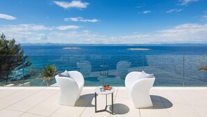 Un balcon avec deux chaises blanches et une petite table, d’où l’on peut admirer une mer bleue et limpide, avec des îles qui se dessinent à l’horizon.