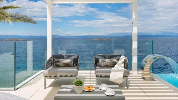 Un balcon avec deux chaises, une table ornée d’une plante et d’une assiette de nourriture, et une balustrade en verre donnant sur la mer.