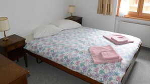 Een slaapkamer met een bloemenbedekking, twee bedlampen, een houten nachtkastje, een houten dressoir en een raam met beige gordijnen.
