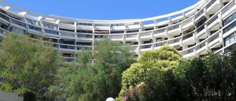 A modern, multi-story building with a curved facade, surrounded by lush greenery and a clear blue sky.