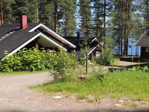 Een chalet met schuin dak en schoorsteen, omgeven door dicht bos en een grindpad.