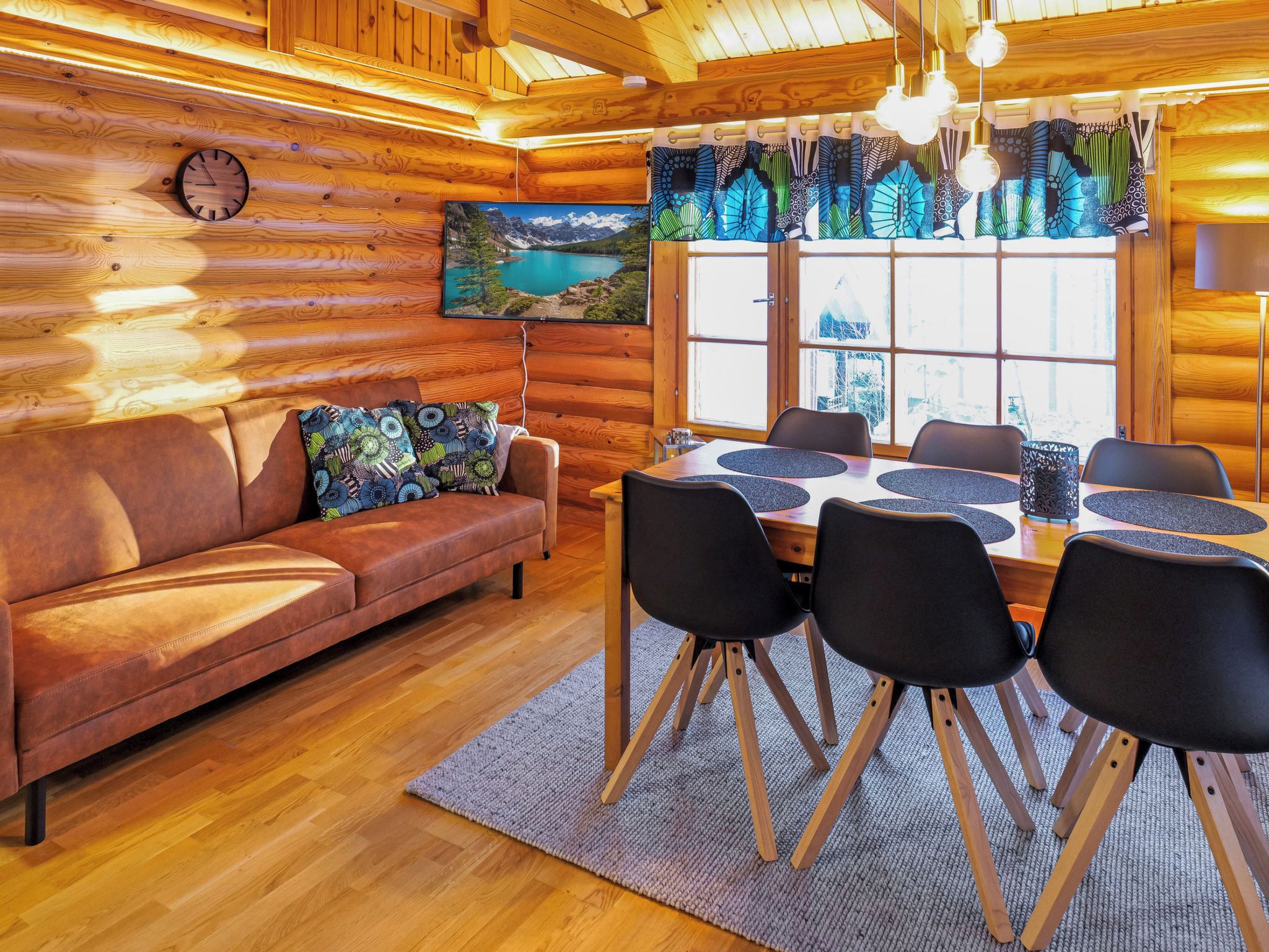Interior acogedor de una cabaña con pared de madera, un sofá con cojines estampados, una mesa de comedor con sillas y un ventanal con cortinas.