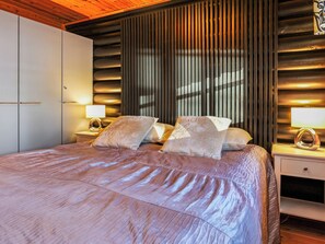 A bedroom with a large bed, two bedside tables with lamps, a wardrobe, and a window with vertical blinds.