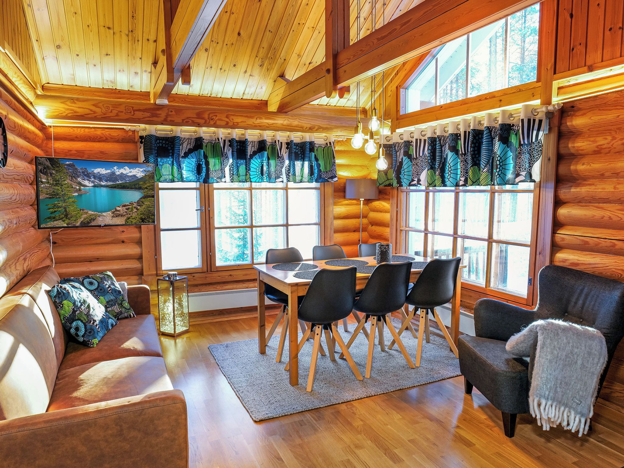 A cozy cabin interior with a wooden ceiling, a dining table with chairs, a sofa, and a large window with curtains.
