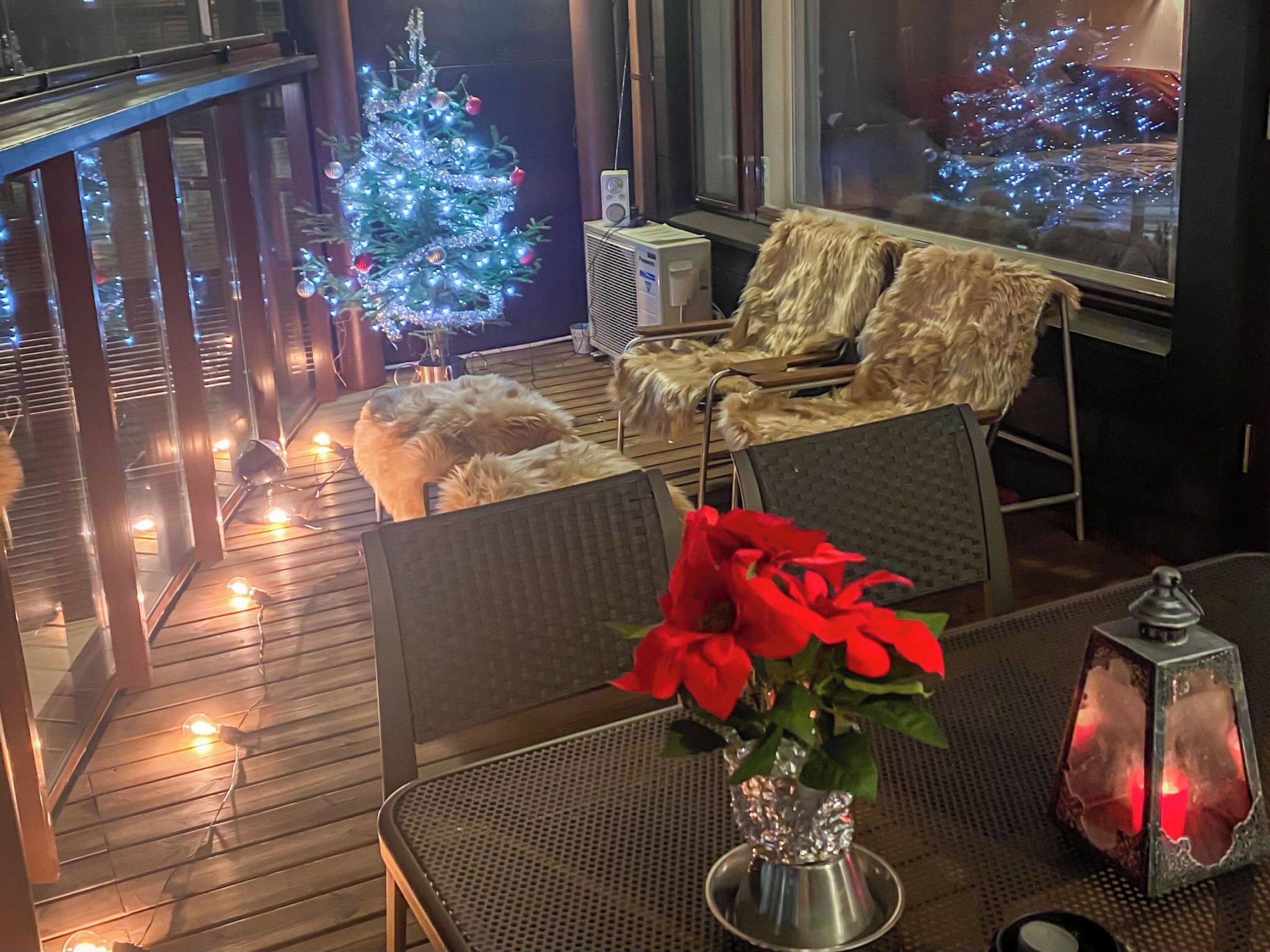 Un espace intérieur chaleureux avec une terrasse en bois, un petit sapin de Noël, un fauteuil recouvert de fourrure et une table ornée d’un vase rempli de fleurs rouges.