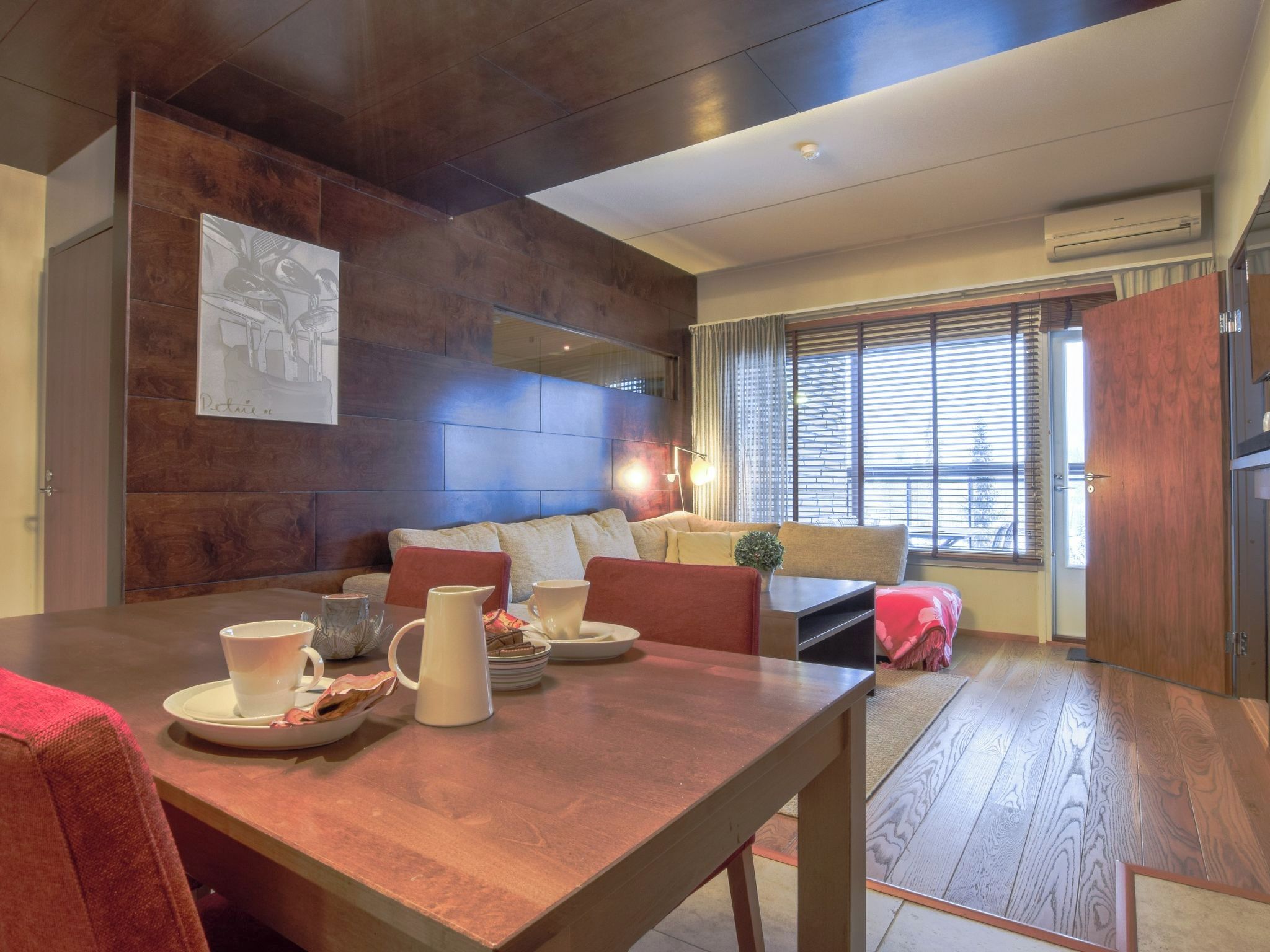 A modern living room with a wooden dining table, a sofa set, and a large window with blinds.