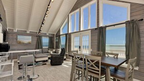 A spacious room with a high ceiling, large windows, a dining area, and a seating area with a view of the sea.