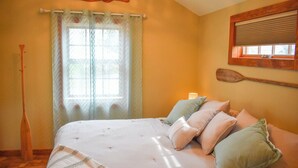 Ein Schlafzimmer mit einem Bett, Kissen, einem Fenster mit Vorhängen, einem Holzschläger und einem an der Wand montierten Fensterladen.