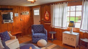 Ein gemütliches Blockhaus-Zimmer mit Holzwänden, einem blauen Sessel mit Anker-Motiv, einem kleinen runden Tisch, einer weißen Kommode mit dekorativen Griffen und einem Fenster mit weißen Vorhängen.
