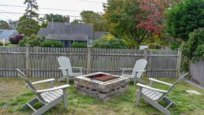 Ein Garten mit einer Holzterrasse, einem Feuerkorb und Adirondack-Stühlen.