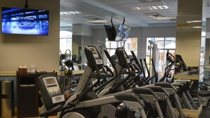 Een rij stilstaande fietsen in een sportschool, met een televisie aan de muur waarop een nieuwskanaal wordt vertoond.
