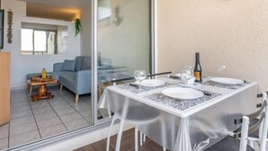 A dining area with a table set for two, a glass partition, and a view of a living room with a sofa and a small table. - Copacabana by Interhome (Canet-Plage)
