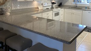 Cocina con gabinetes blancos, encimera de granito, microondas y estufa. Hay tres taburetes de bar y luces colgantes.