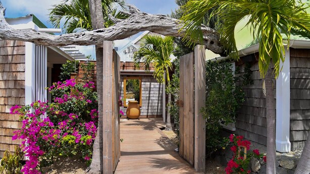 Un sendero de madera lleva a una casa con un arco rústico, rodeada de coloridas flores de buganvilla y palmeras.