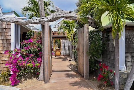 A wooden pathway leading to a house with a rustic archway, surrounded by vibrant bougainvillea flowers and palm trees.