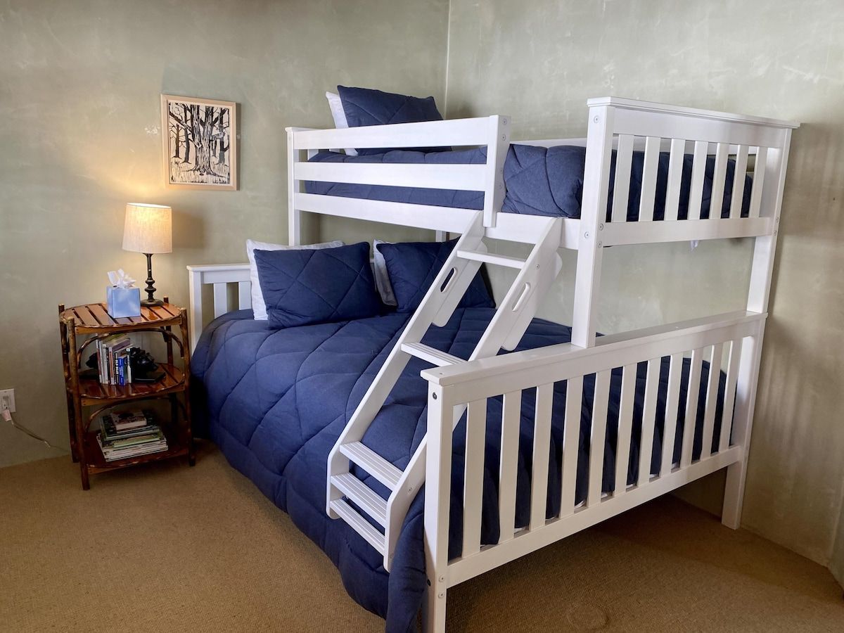 A bunk bed with a blue bedspread and white railings, a small wooden nightstand with books and a lamp, and a framed picture on the wall.