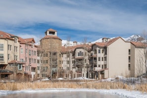 Un hôtel à plusieurs étages, doté d’une tour centrale, entouré de bâtiments avec des balcons et surplombant un paysage enneigé.