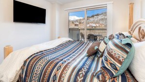 A bedroom with a large bed, a flat-screen TV mounted on the wall, and a sliding glass door leading to a balcony with a view of a snowy landscape.
