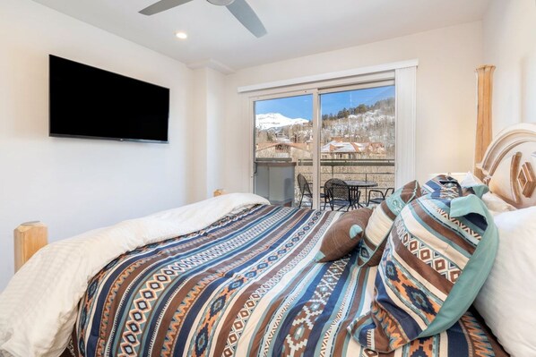 A bedroom with a large bed, a flat-screen TV mounted on the wall, and a sliding glass door leading to a balcony with a view of a snowy landscape.