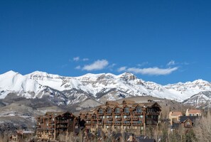 A multi-story lodge with a complex architectural design, situated in a mountainous area with snow-covered peaks in the background.