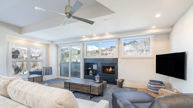A modern living room with a fireplace, large windows, and a ceiling fan. - Penthouse At The Peaks 751 (Mountain Village)