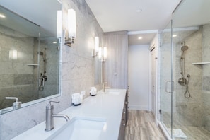 Un baño moderno con ducha envuelta en vidrio, un espejo grande y un mostrador blanco con dos lavamanos.