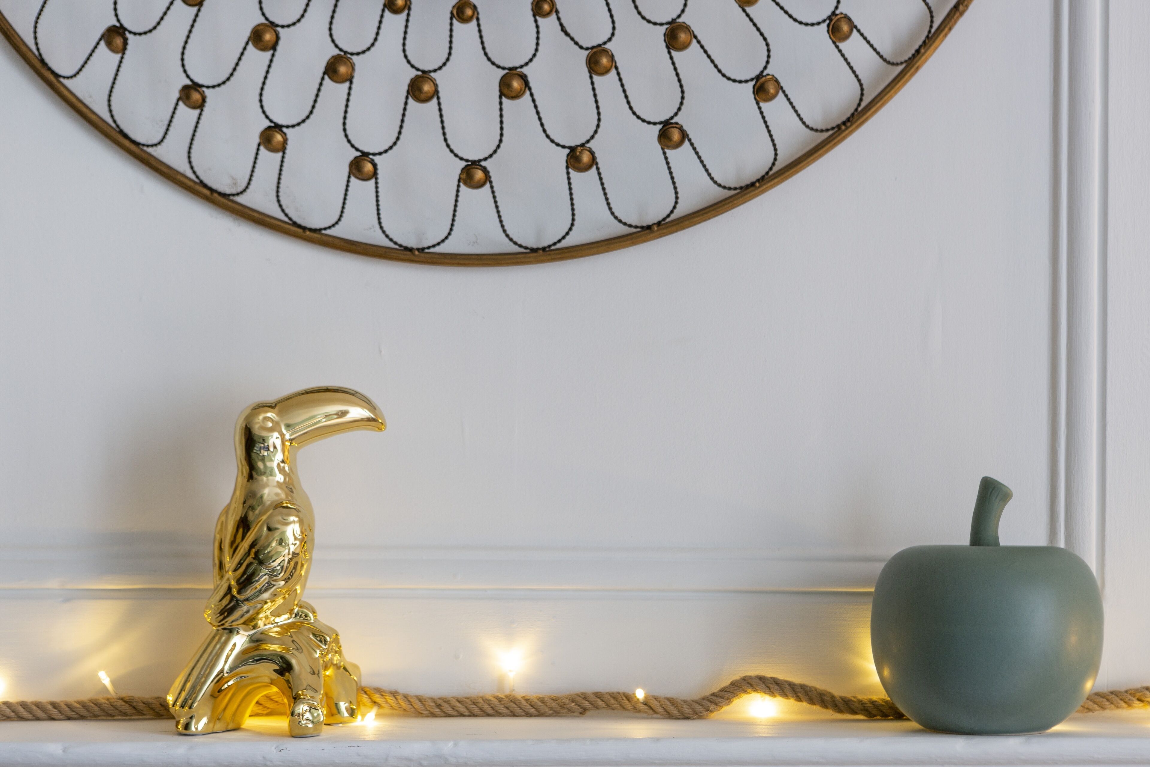 Una pared decorativa con un diseño metálico estampado, una escultura de pájaro dorado y una manzana verde ornamental sobre una repisa blanca.