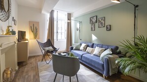 A living room with a blue sofa, a grey armchair, a coffee table, and a floor lamp. There is a cactus painting on the wall and a large plant in the corner. - LE TOUCAN, 2 pièces de charme et emplacement de rêve - Parlement de Bretagne (Rennes)