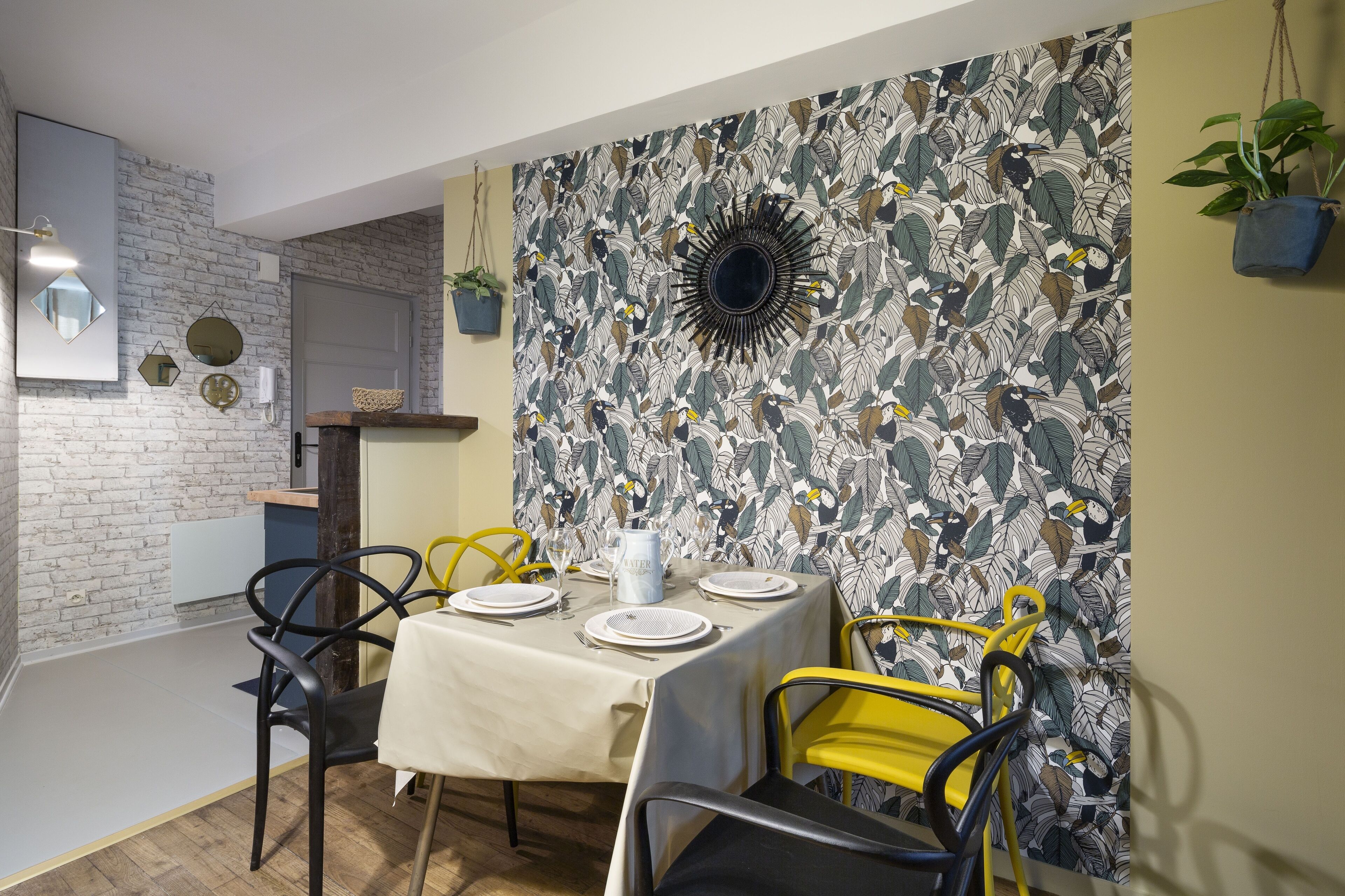 A dining area with a brick wall, a floral wallpaper, and a sunburst mirror. There are yellow chairs and a table set with plates and a pitcher.