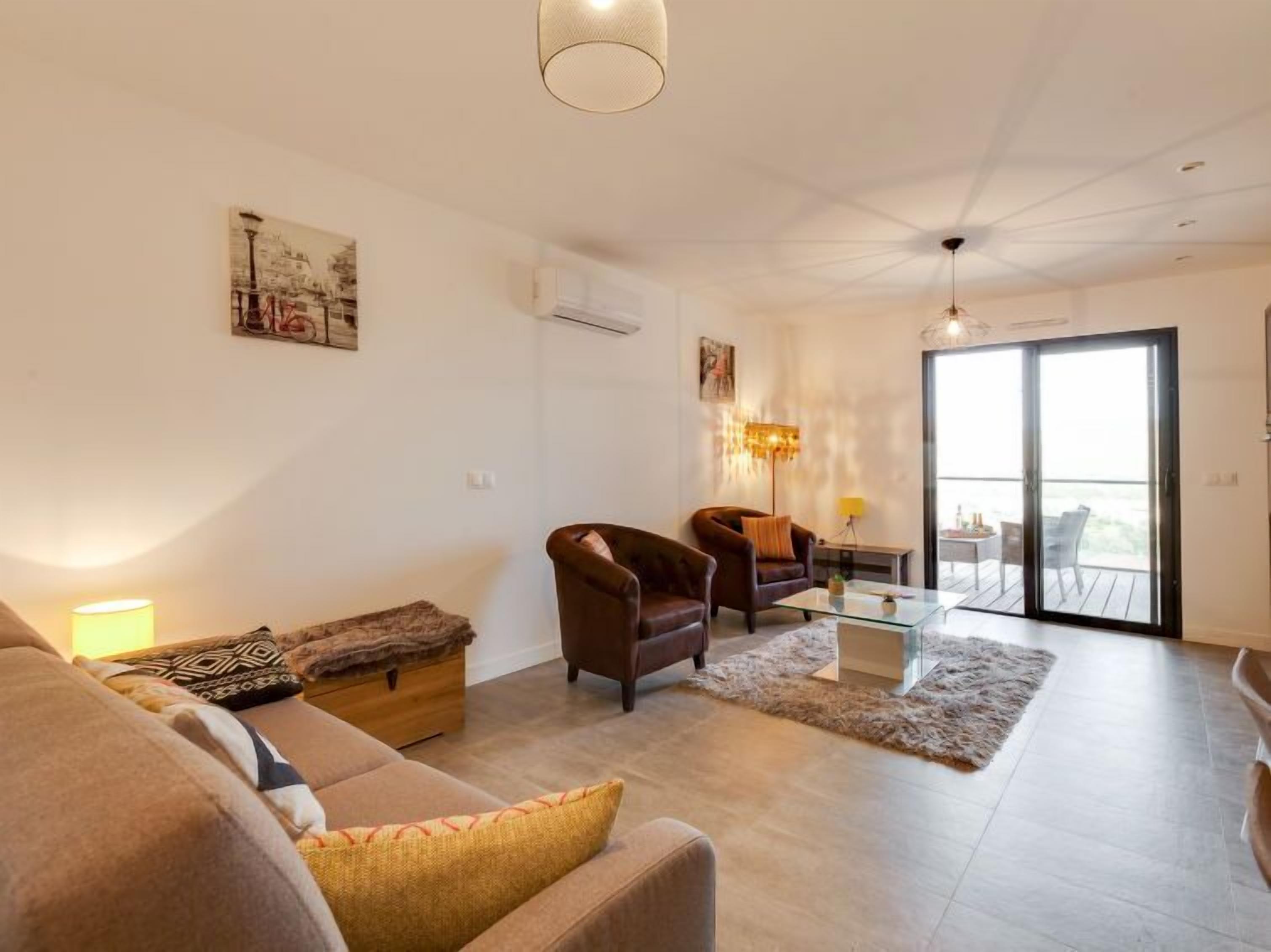 Un salon moderne avec un canapé, des fauteuils et une table basse en verre. Une porte-fenêtre coulissante donne accès à un balcon extérieur.