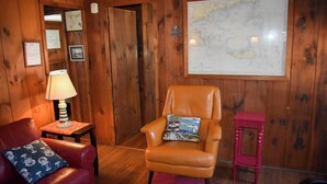 Une pièce chaleureuse aux murs en bois, ornée d’une grande carte, avec un canapé en cuir rouge, un fauteuil beige et un petit tabouret rouge.