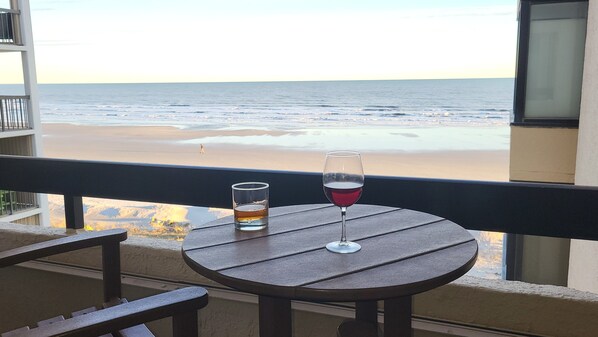 A balcony with a table, a glass of red wine, and a glass of beer, overlooking a beach and ocean.