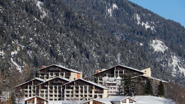 Une station de ski comprenant plusieurs bâtiments de style chalet, une piste de ski et un paysage montagneux en toile de fond.