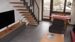 Un salon moderne avec un téléviseur à écran plat, un escalier en bois, un canapé gris et un coin repas avec une nappe rouge.