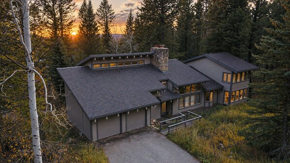 A modern two-story house with a stone chimney, large windows, and a driveway leading to a gated entrance.