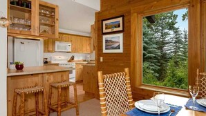 Interno di una baita in legno con cucina, zona pranzo e ampie finestre che si affacciano su una foresta.