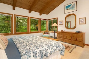 A bedroom with a large bed, a wooden dresser, and a mirror. There are multiple windows providing a view of greenery outside.