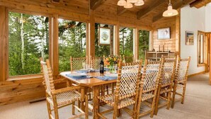 Zona pranzo in legno con tavolo imbandito per sei persone, circondata da sedie in vimini, ampie finestre con vista sul bosco e lampadario a sospensione.