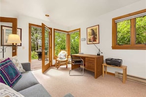 A room with a desk, chair, and a view of greenery through large windows.