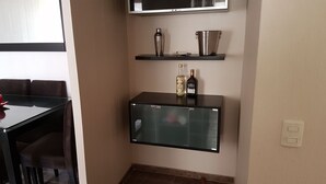 A small kitchenette with a glass-fronted cabinet, a shelf with a bottle and a container, and a chair at a table.