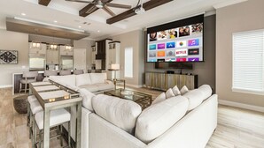 A modern living room with a large flat-screen TV, a ceiling fan, and a dining area with a glass coffee table. - Reunion Fantasy | Games Room, Secret Playroom, English Pub Bar (Kissimmee)