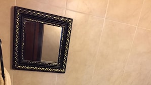Ein Badezimmer mit einem beigen Waschbecken, schwarzen Wasserhähnen, einem Spiegel mit goldener Rahmung, einer Toilette und einer gefliesten Rückwand mit Muster.