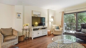 Ein Wohnzimmer mit Fernseher, zwei Sofas, einem Glastisch und einem Balkon mit Blick auf Grünflächen.