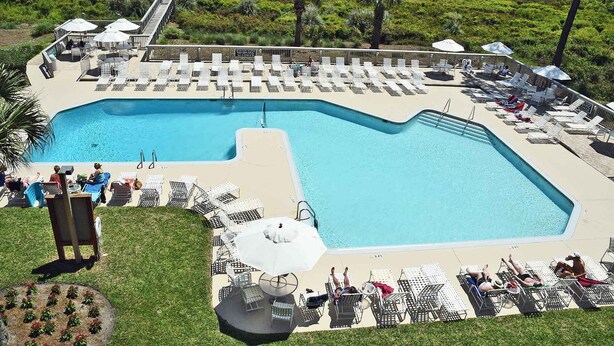 Ein großer Pool mit Liegestühlen und Sonnenschirmen, umgeben von Grünflächen und Blick auf einen Strand.