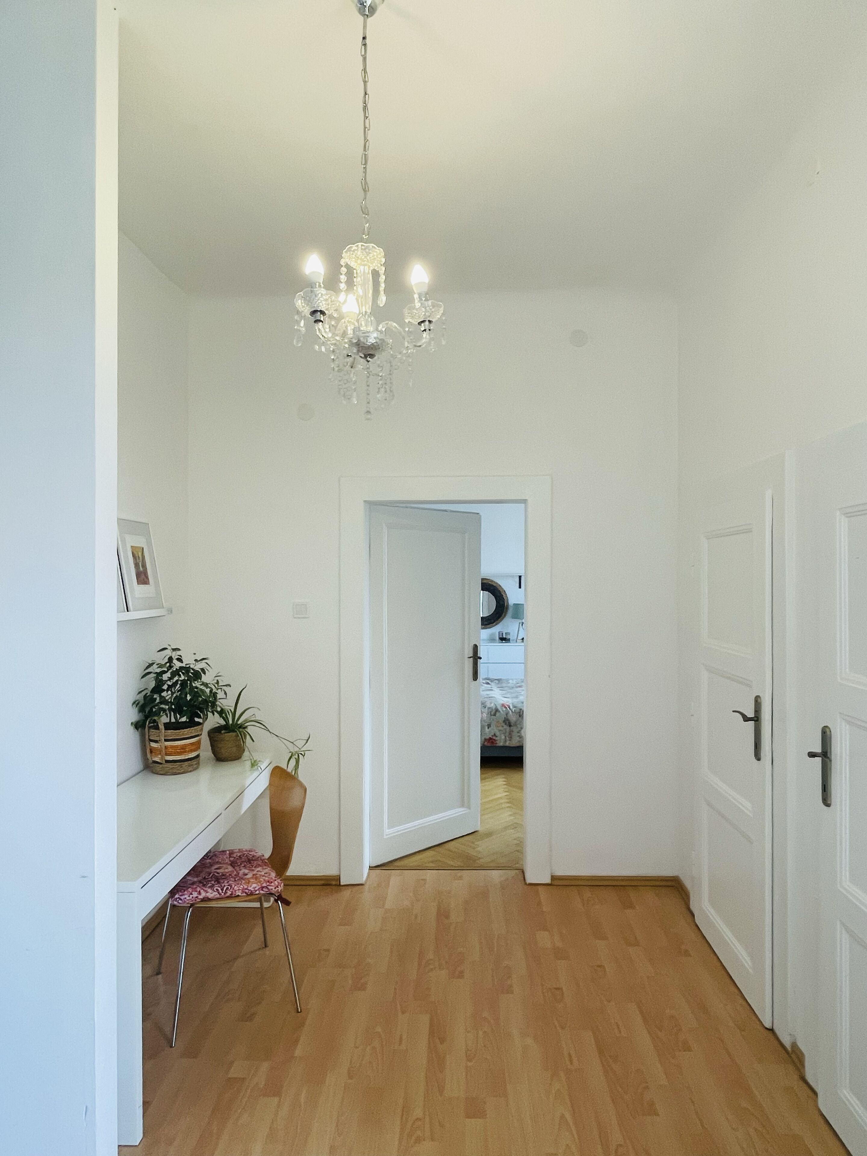 Ein Flur mit Holzboden, weißen Wänden, einer Chandelierleuchtre, einem Schreibtisch mit Stuhl und einer Tür, die in einen anderen Raum führt.