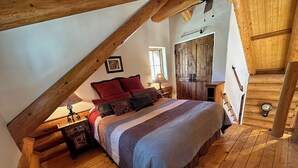 Un dormitorio acogedor con techo inclinado, suelo de madera, una cama grande con muchas almohadas, una mesita de noche, un aparador y un armario de madera.