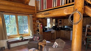 Interior acogedor de una cabaña con sala de estar, muebles de madera, una cocina y un amplio ventanal con vistas a un bosque.